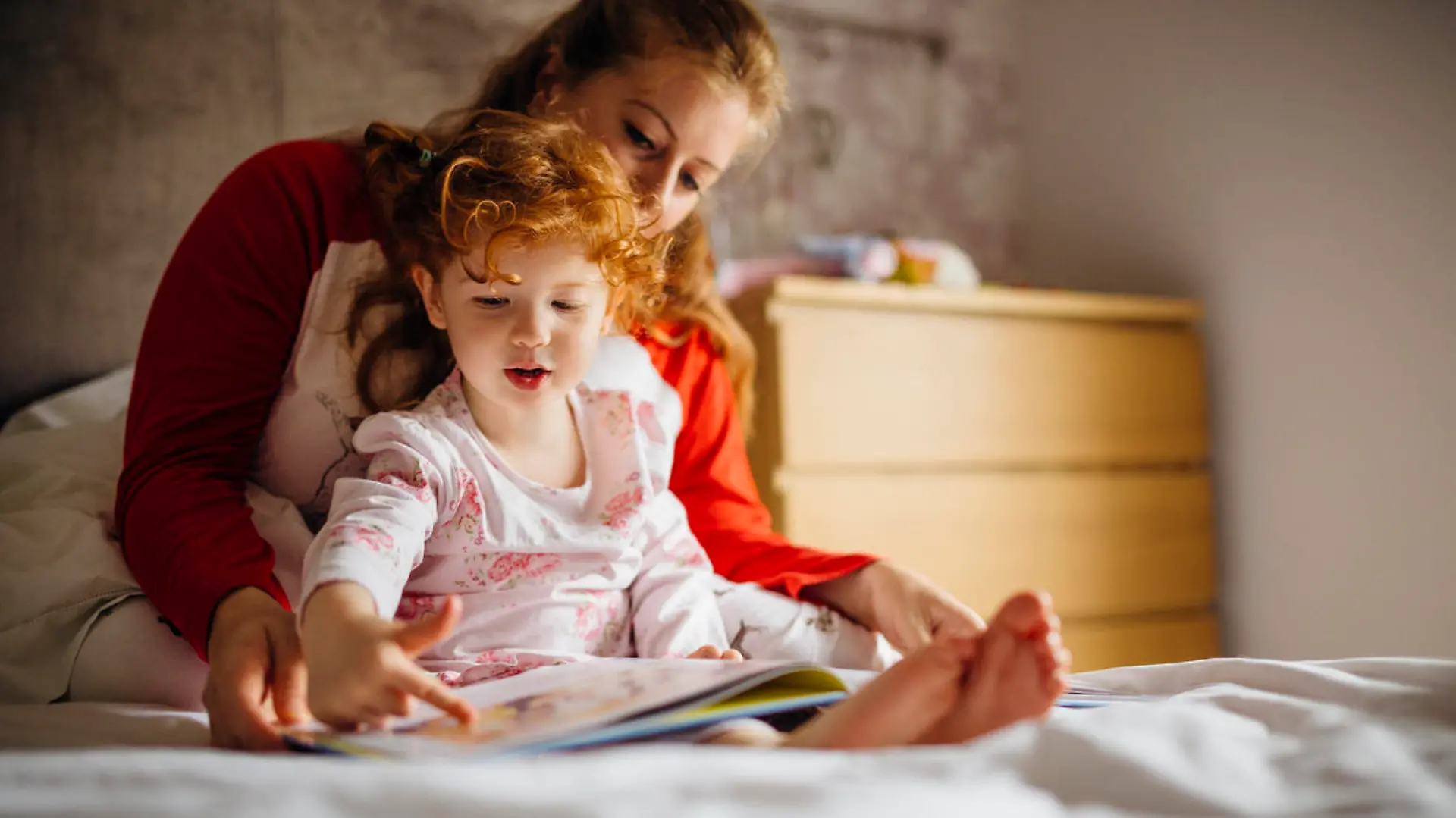 Little girl reading a bedtime story with her Mother in her bed. They are sitting together and the little girl is pointing at one of the pictures.