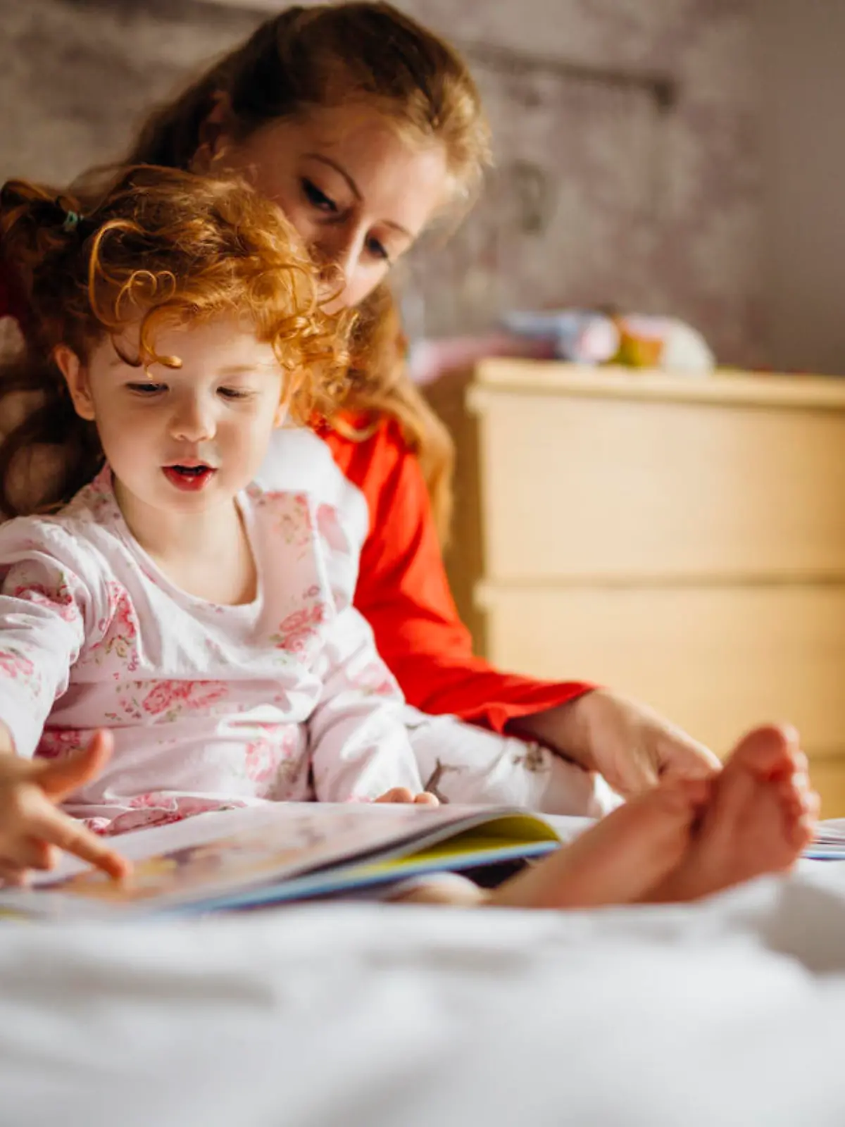 Little girl reading a bedtime story with her Mother in her bed. They are sitting together and the little girl is pointing at one of the pictures.