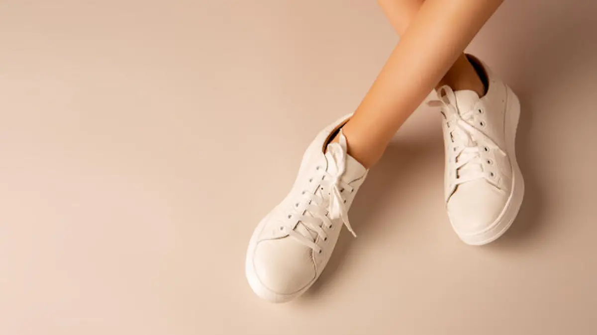 Casual footwear - white leather sneakers shoes and girlâs crossed legs on nude background. Layout with free copy (text) space.