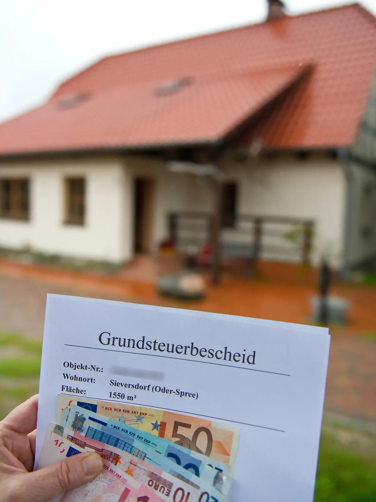 ARCHIV - 09.09.2010, Brandenburg, Sieversdorf: ILLUSTRATION - Eine Frau hält  Euro-Geldscheine und einen Grundsteuerbescheid vor ihrem Haus im brandenburgischen Sieversdorf (Oder-Spree) in der Hand (Illustration zum Thema Grundsteuer). Bis Ende Juni seien bei den Finanzämtern rund 1 052 000 Erklärungen eingegangen, teilte das Justizministerium auf Anfrage mit. Damit fehlten immer noch knapp 200 000 Grundsteuererklärungen. (zu dpa: «Grundsteuer: 200 000 Immobilienbesitzern droht Strafzahlung») Foto: Patrick Pleul/dpa-Zentralbild/dpa +++ dpa-Bildfunk +++