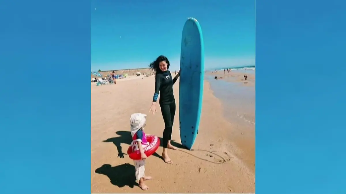 Rebecca Mir posiert mit einen Surfbrett unter dem Arm mit ihrem kleinen Sohn am Strand.