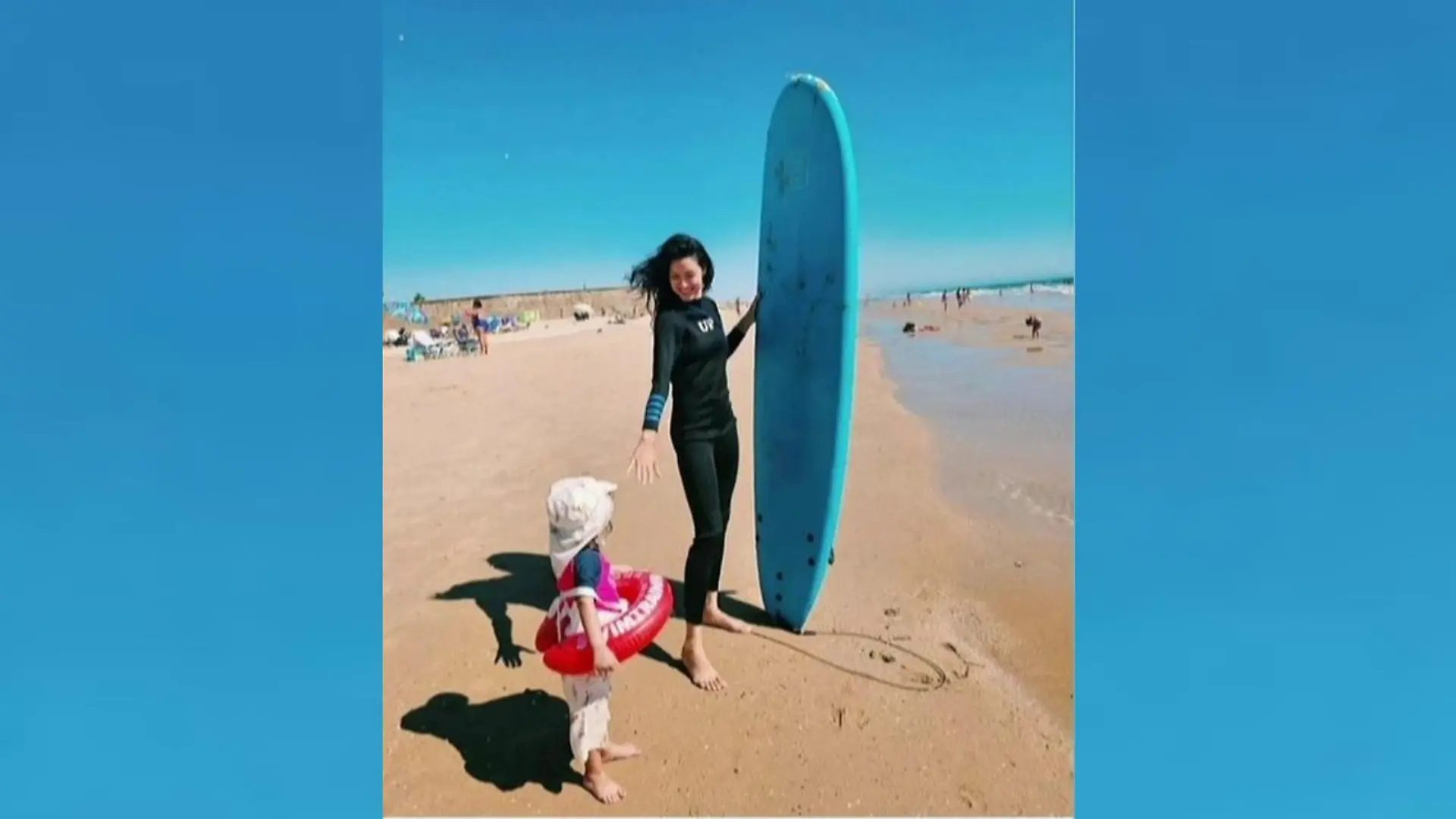 Rebecca Mir posiert mit einen Surfbrett unter dem Arm mit ihrem kleinen Sohn am Strand.