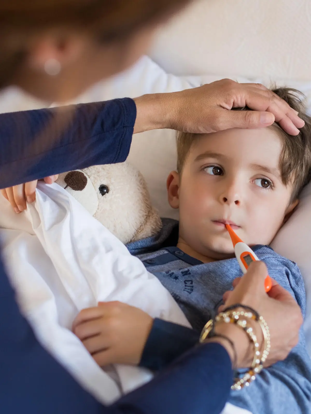 Sick boy with thermometer laying in bed and mother hand taking temperature. Mother checking temperature of her sick son who has thermometer in his mouth. Sick child with fever and illness while resting in bed.