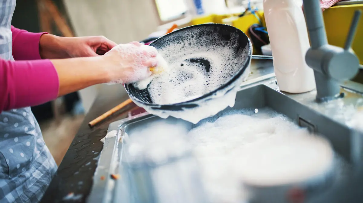 Closeup of unrecognizable woman doing dishes with lots of dirty plates and pots waiting aside. She's cleaning a black non stick cooking pan.