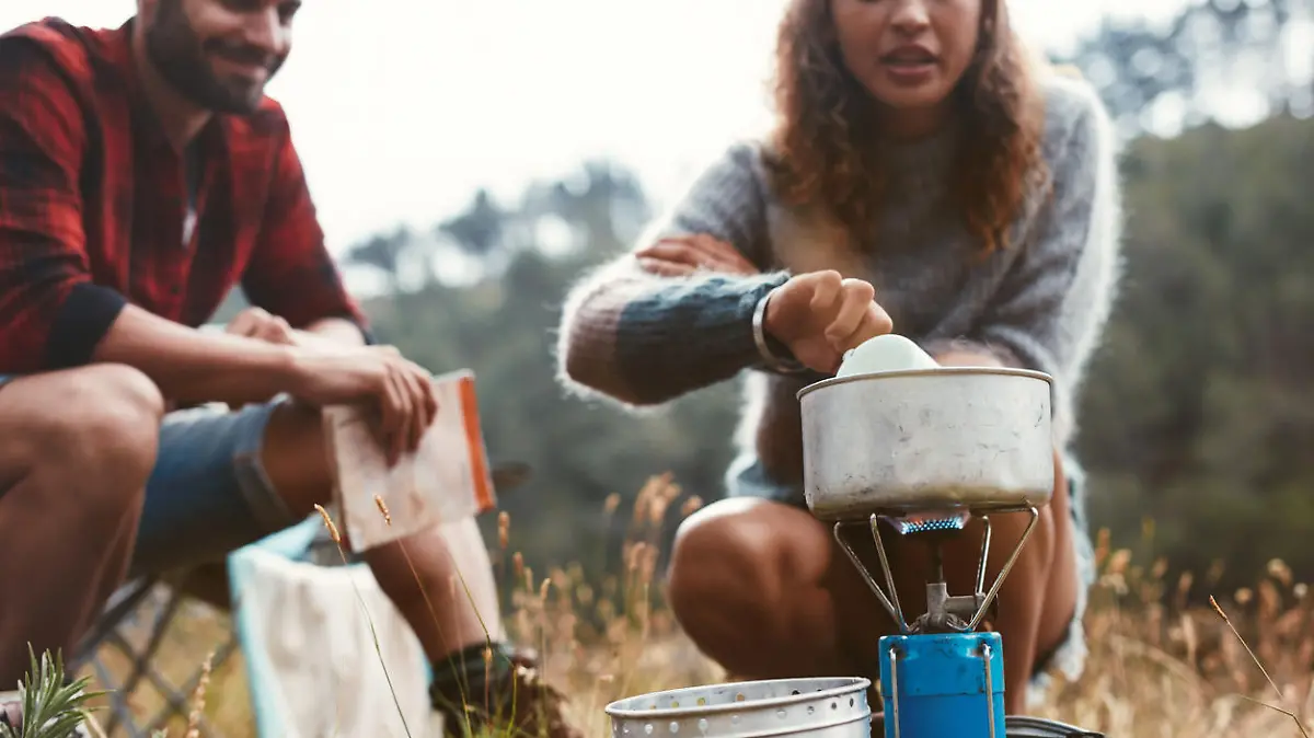 Young woman preparing food on a gas stove while camping with her boyfriend. Adventurous young couple camping in the forest.