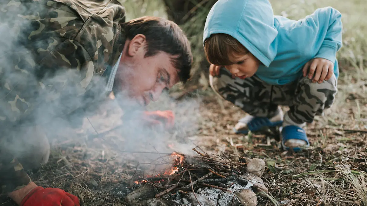 little happy five year old kid boy and his father blowing and kindles a campfire in the village or forest on nature outdoor on a summer evening. authentic countryside and children's rustic life