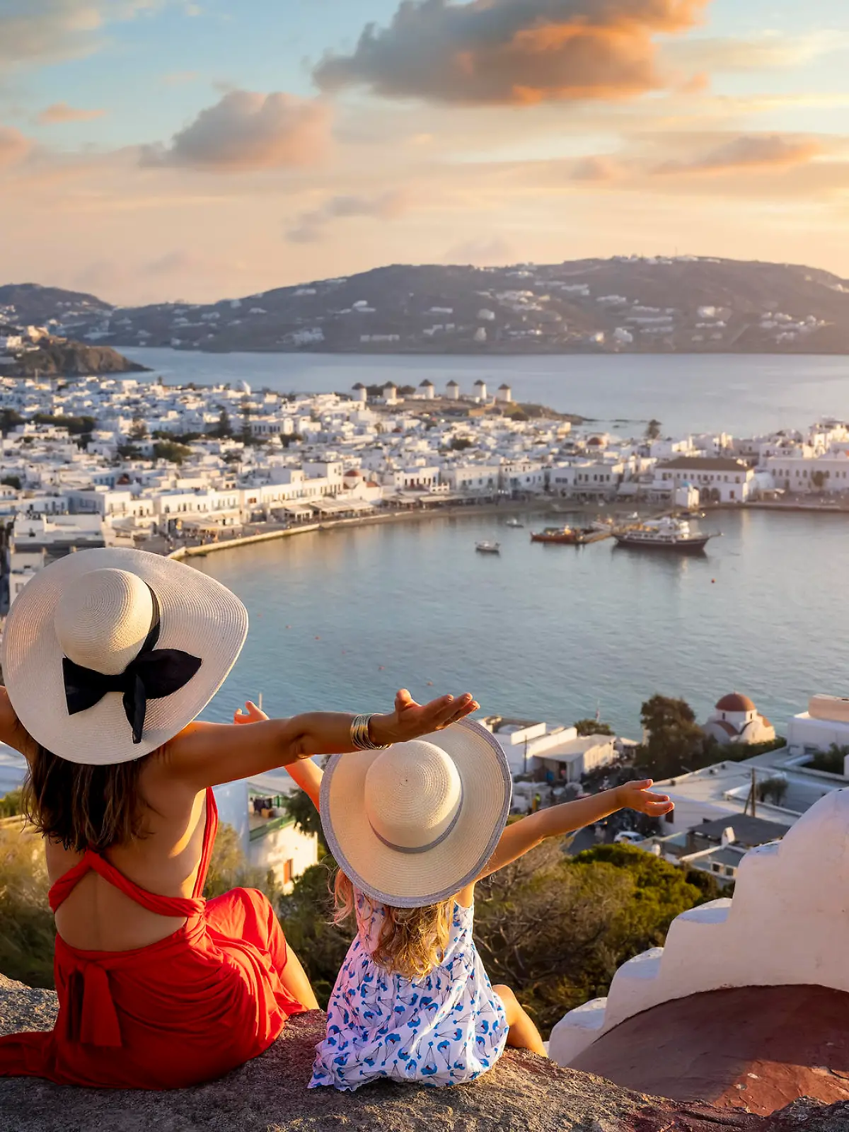 A happy mother and daughter on family holidays overlook the town of Mykonos island during a summer sunset, Cyclades, Greece