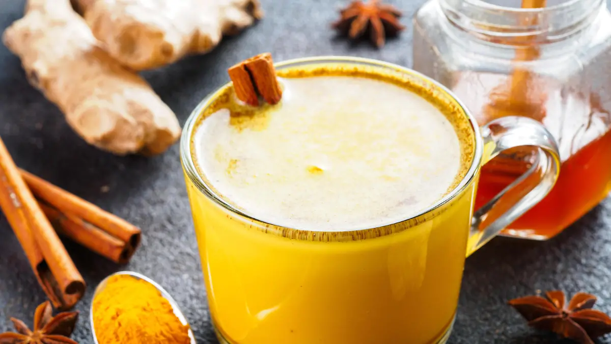 Healthy drink golden turmeric latte in glass cup.Gold milk with turmeric,ginger root,cinnamon sticks,dried turmeric powder and honey over black cement background.Detox turmeric tea and ingredients.