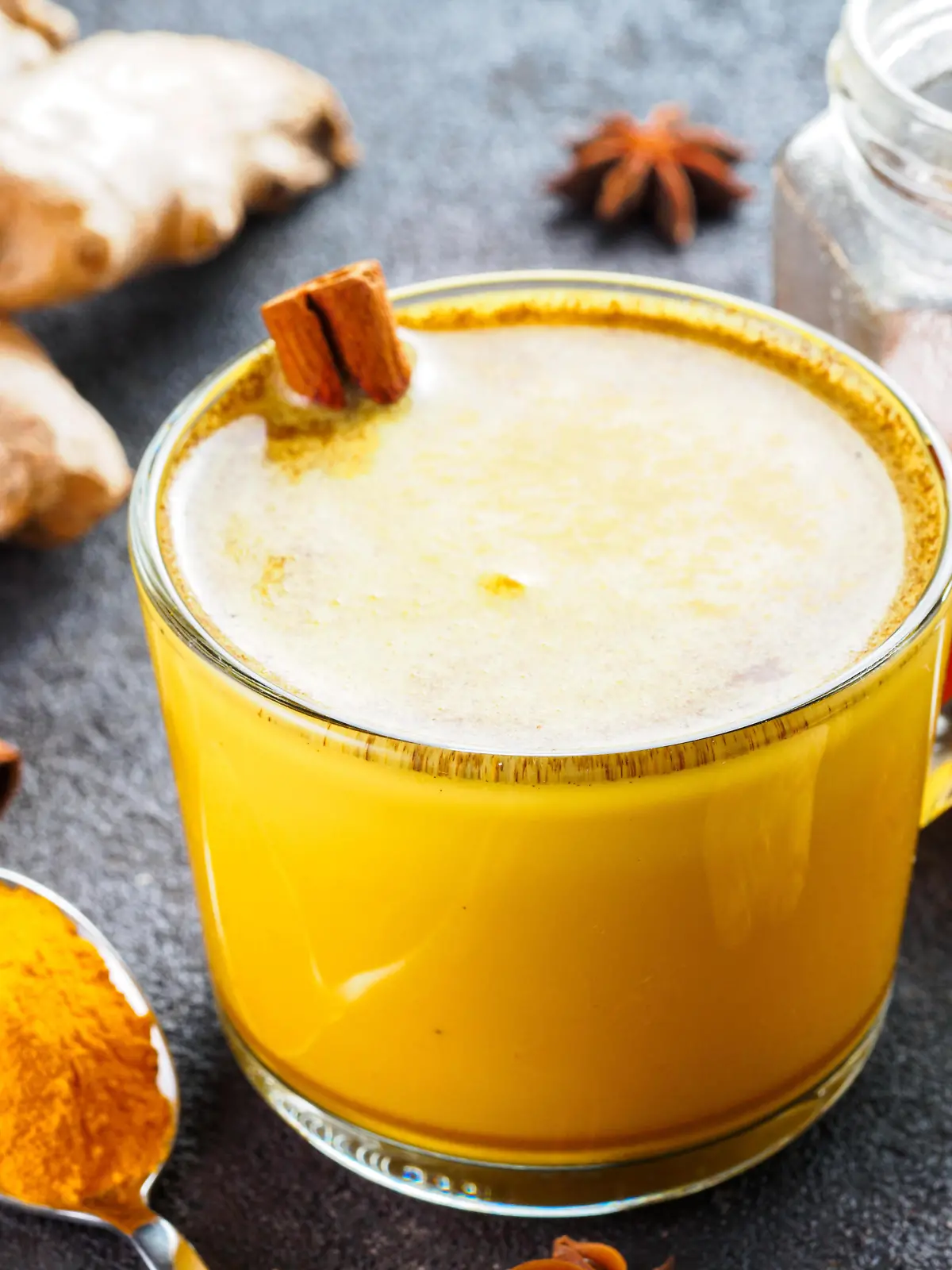 Healthy drink golden turmeric latte in glass cup.Gold milk with turmeric,ginger root,cinnamon sticks,dried turmeric powder and honey over black cement background.Detox turmeric tea and ingredients.