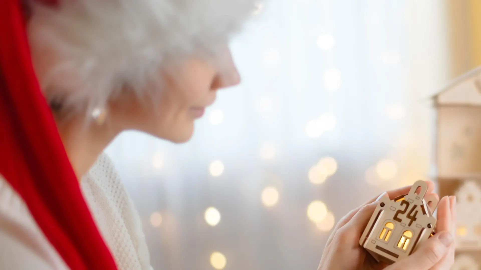 The girl's hands hold out a small wooden house with the number 24 on the background of the Advent calendar with yellow bokeh lights. The atmosphere of celebration, bokeh light.