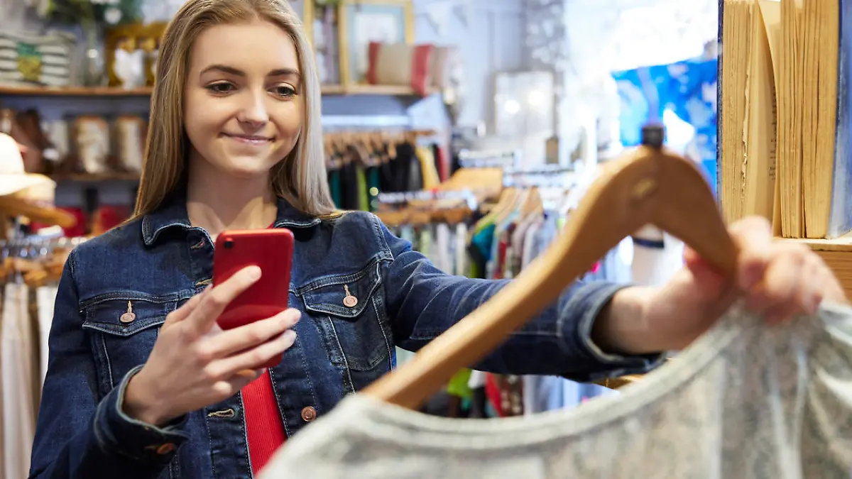 Young Woman Buying Used Sustainable Clothes From Second Hand Charity Shop Taking Picture Of Dress On Mobile Phone