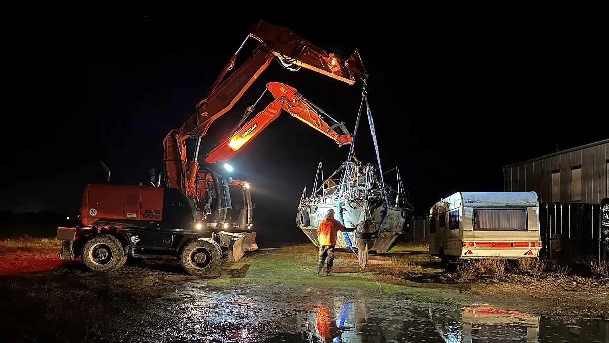 15.12.2023, Niedersachsen, Norderney: Zwei Bagger heben das im Hafen der Insel Norderney abgestellte und Anfang November gestrandete Segelboot für einen geplanten Abtransport zum Festland an. Ein für den Freitagmorgen geplanter Abtransport ans Festland mit einem Tieflader und einem Frachtschiff kam allerdings nicht zustande. (zu dpa: "Auf Norderney gestrandetes Segelboot kommt nicht von Insel los") Foto: Volker Bartels/dpa +++ dpa-Bildfunk +++