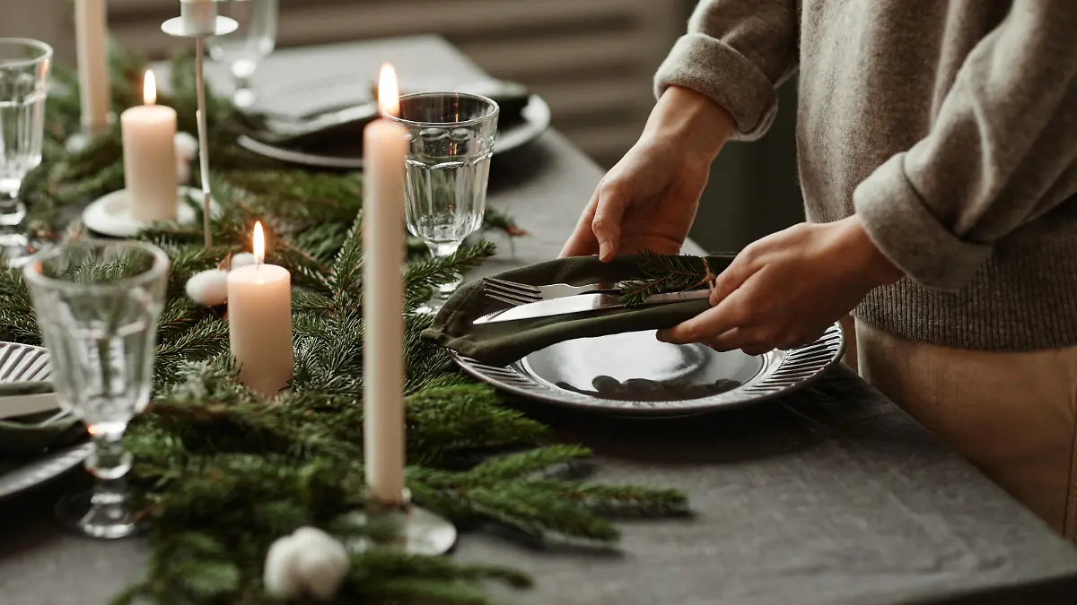 Side view close up of unrecognizable woman setting up dining table decorated for Christmas with fir branches and candles in grey tones, copy space