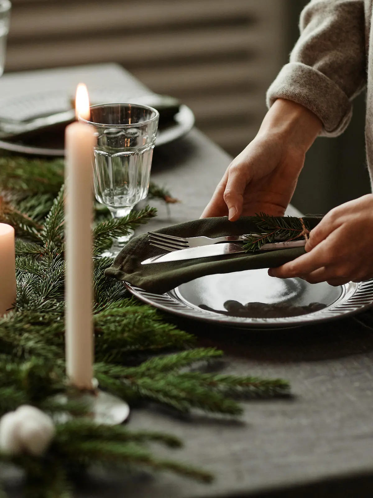 Side view close up of unrecognizable woman setting up dining table decorated for Christmas with fir branches and candles in grey tones, copy space