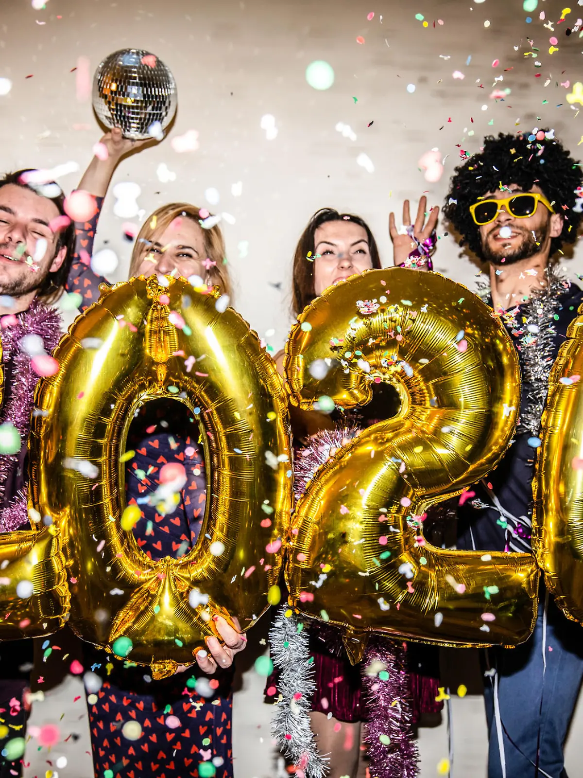 Four friends are celebrating the New Year 2024 with a party and big shiny foil balloons for 2024 and lots of falling confetti