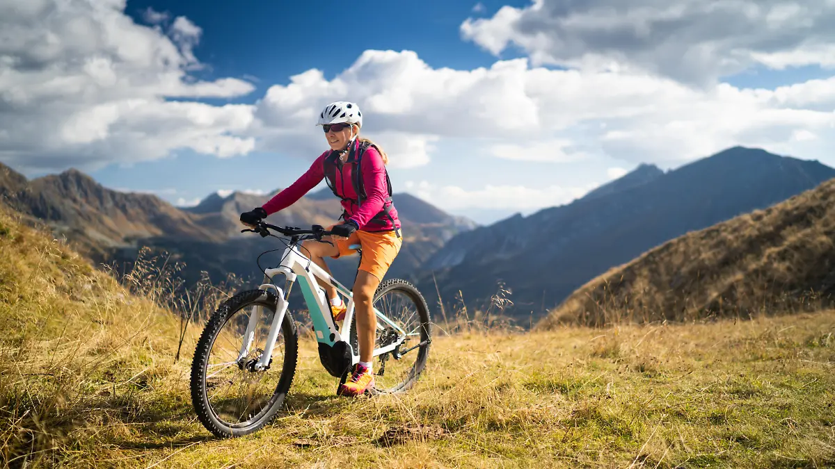 smiling fit sporty woman cycling her electric mountain bike high up in mountains on sunny autumn day with white clouds