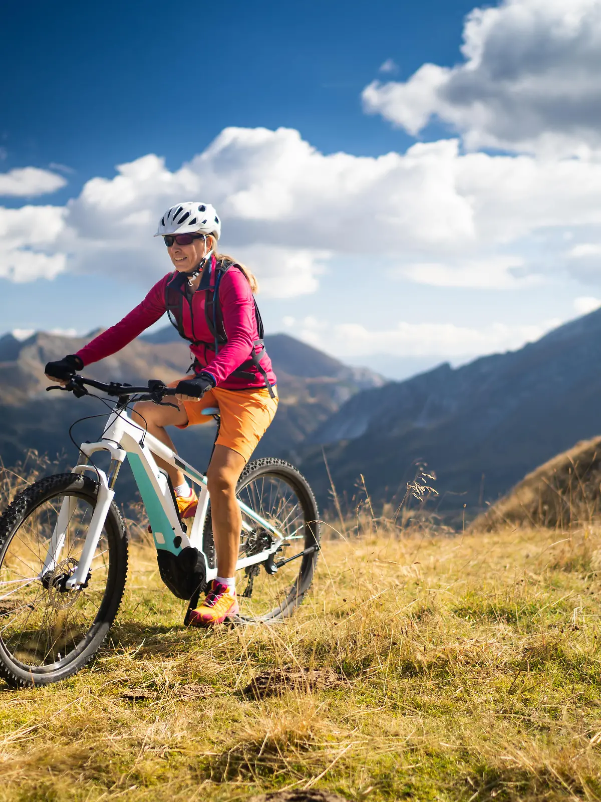 smiling fit sporty woman cycling her electric mountain bike high up in mountains on sunny autumn day with white clouds