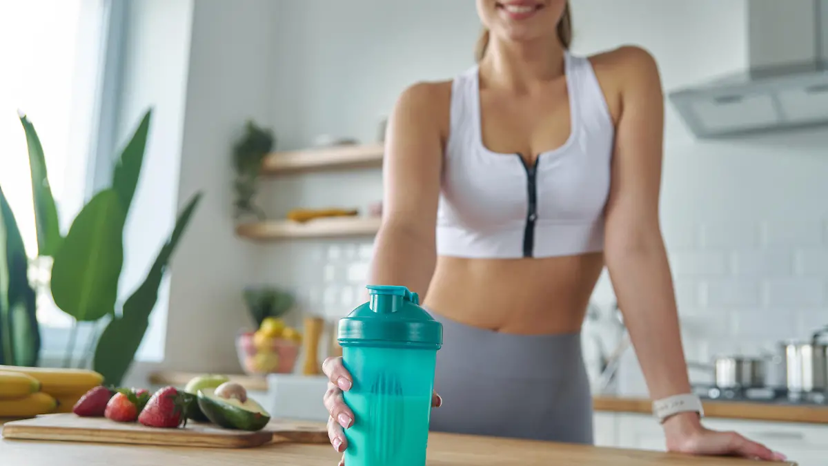 Close-up of woman in sports clothing holding shaker with protein cocktail at the kitchen