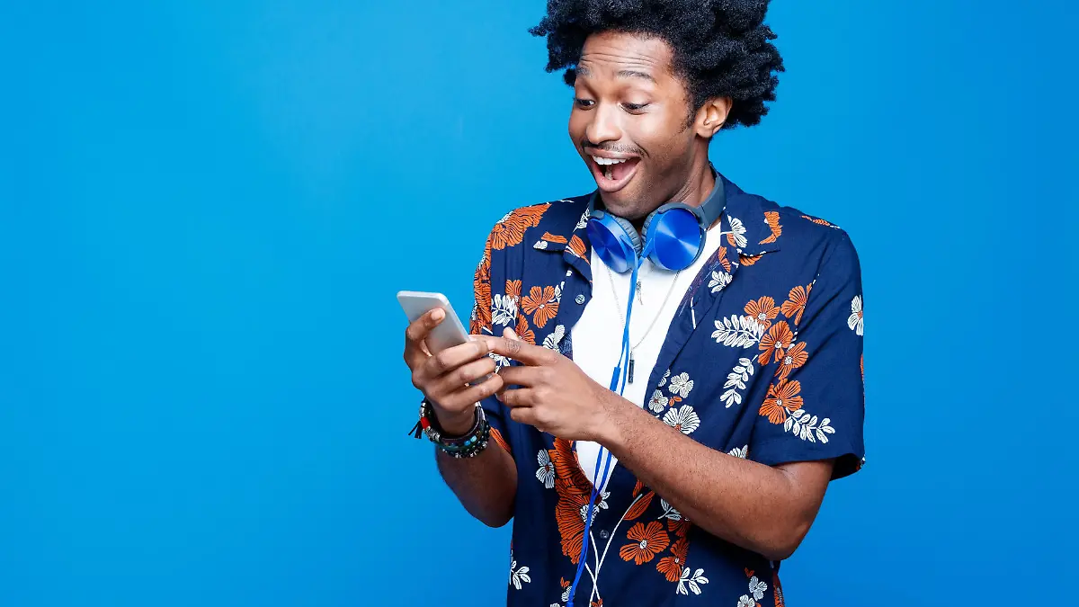Summer portrait of excited afro american young man with dreadlocks wearing hawaiian short sleeved shirt and headphone, holding mobile phone in hands. Studio shot on blue background.