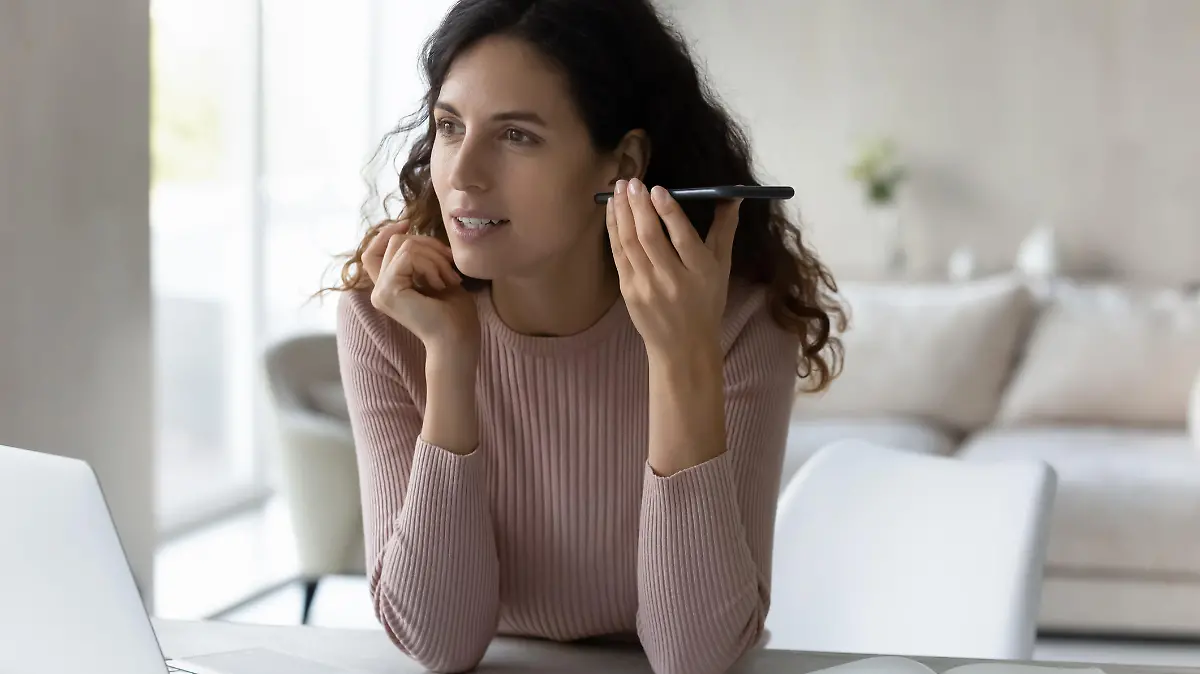 Concentrated young hispanic business woman listening audio message on smartphone, sitting at table with computer. Focused millennial female employee worker communicating distantly using mobile apps.