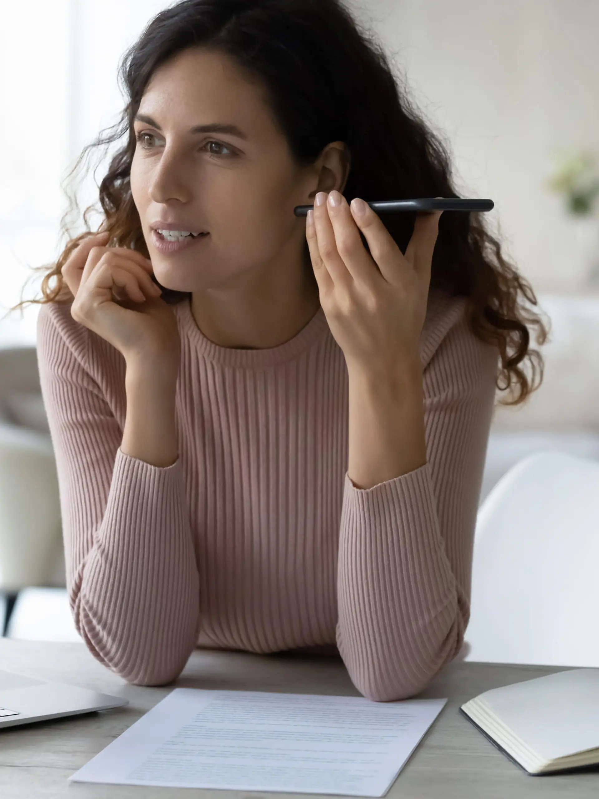 Concentrated young hispanic business woman listening audio message on smartphone, sitting at table with computer. Focused millennial female employee worker communicating distantly using mobile apps.