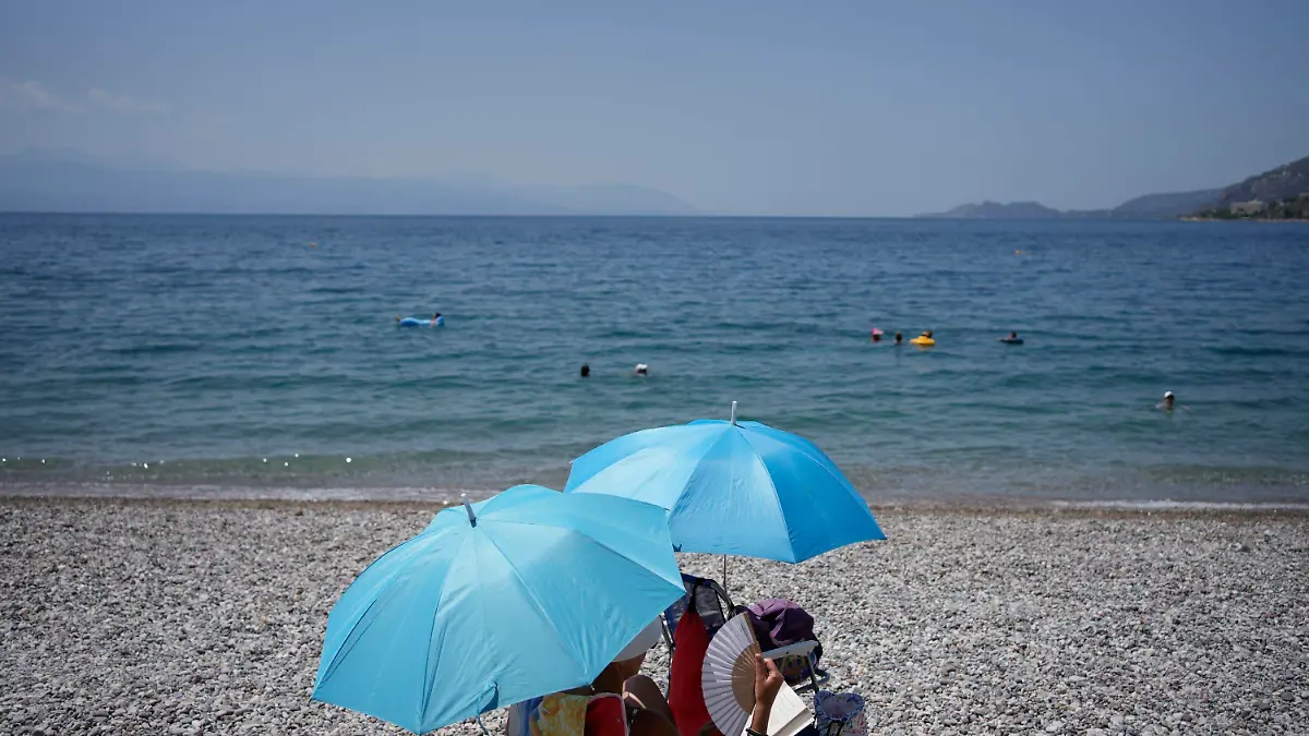 20.07.2023, Griechenland, Loutraki: Eine Frau mit einem Handventilator sitzt unter einem Sonnenschirm am Strand. Es wird erwartet, dass die Temperaturen in Griechenland in den nächsten Tagen noch weiter steigen. Foto: Thanassis Stavrakis/AP/dpa +++ dpa-Bildfunk +++
