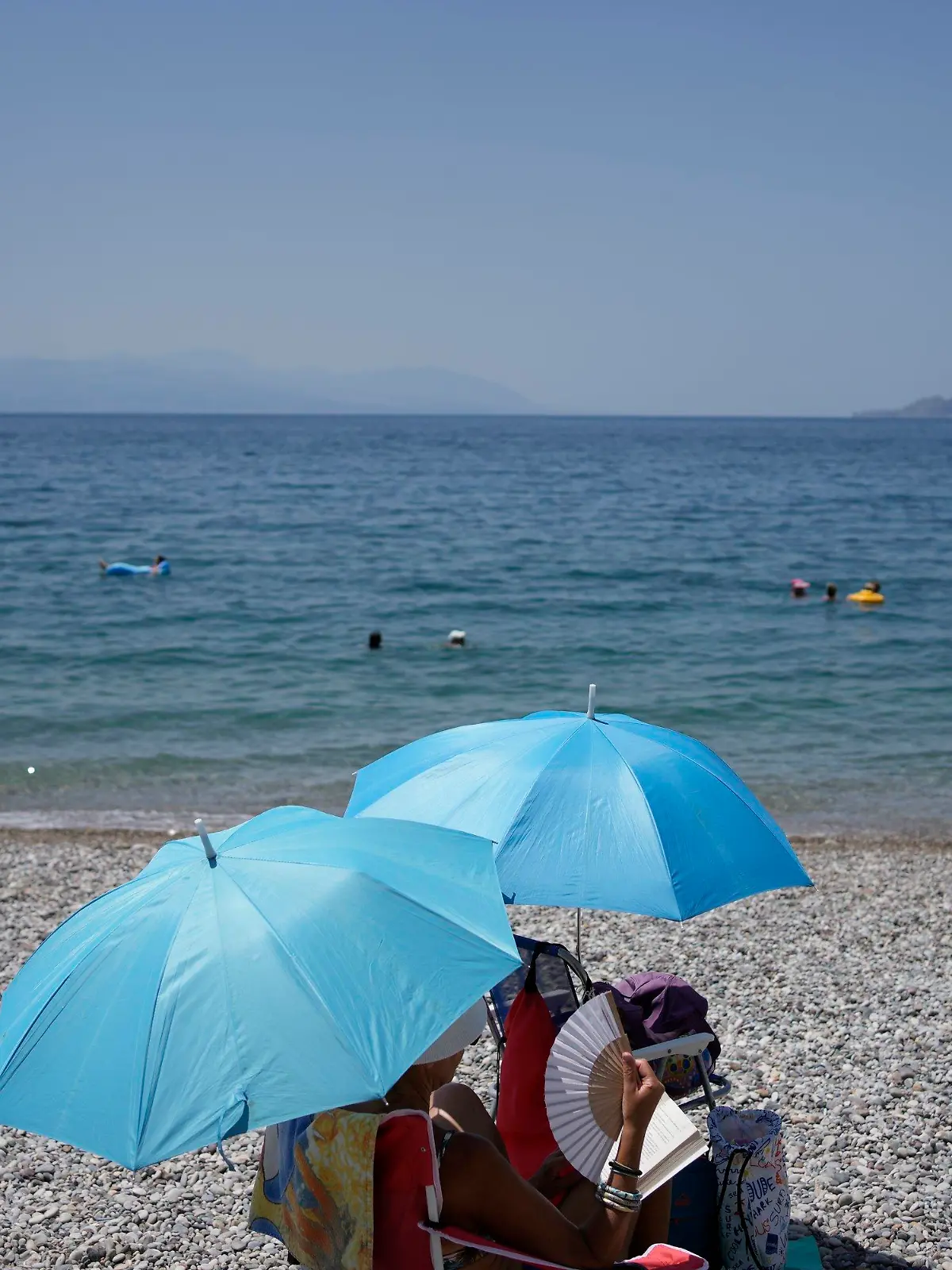 20.07.2023, Griechenland, Loutraki: Eine Frau mit einem Handventilator sitzt unter einem Sonnenschirm am Strand. Es wird erwartet, dass die Temperaturen in Griechenland in den nächsten Tagen noch weiter steigen. Foto: Thanassis Stavrakis/AP/dpa +++ dpa-Bildfunk +++