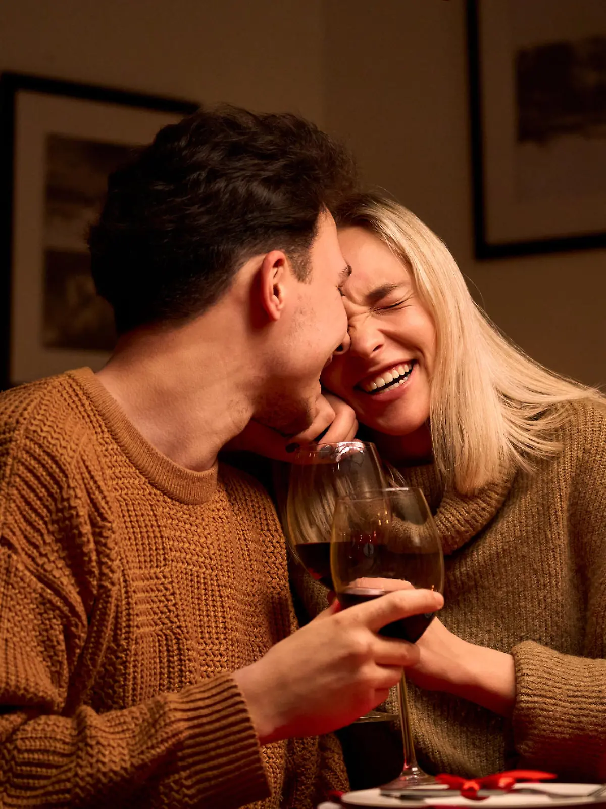 Happy young couple in love hugging, laughing, drinking wine, enjoying talking, having fun together celebrating Valentines day dining at home, having romantic dinner date with candles sitting at table.