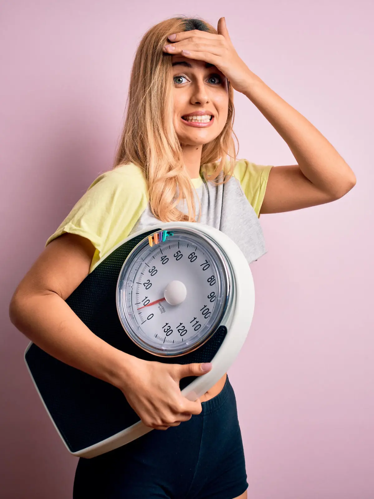 Young beautiful blonde sporty woman on diet holding weight machine over pink background stressed with hand on head, shocked with shame and surprise face, angry and frustrated. Fear and upset for mistake.