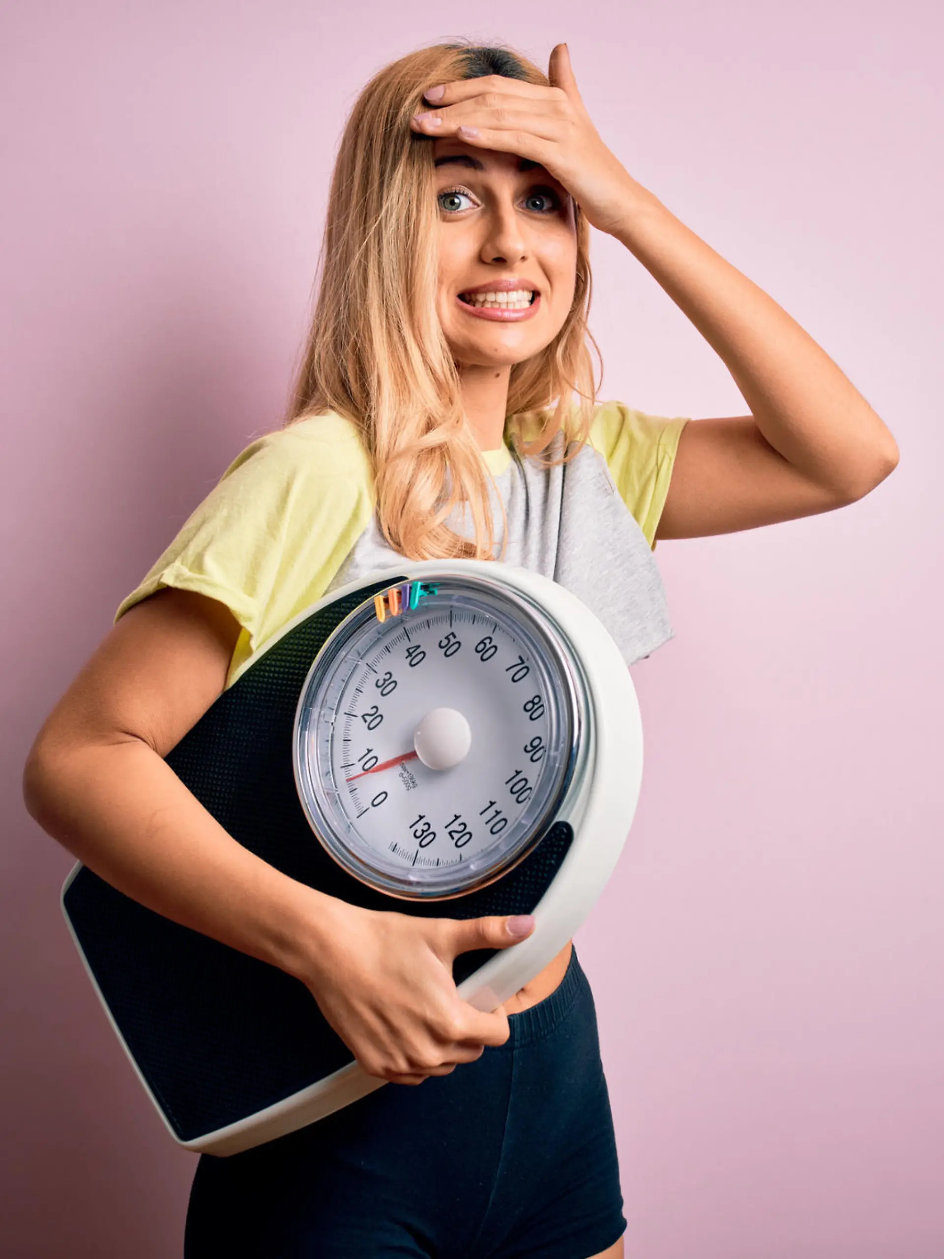 Young beautiful blonde sporty woman on diet holding weight machine over pink background stressed with hand on head, shocked with shame and surprise face, angry and frustrated. Fear and upset for mistake.