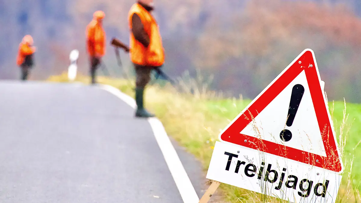 ARCHIV - 20.11.2021, Nordrhein-Westfalen, Wickede: Ein Warnhinweisschild steht während einer Treibjagd am Rande einer Straße. Im Hintergrund stehen Jäger mit Gewehren. (zu dpa: "Jäger erwarten wieder mehr Wildschwein-Abschüsse") Foto: Andreas Dunker/dpa +++ dpa-Bildfunk +++