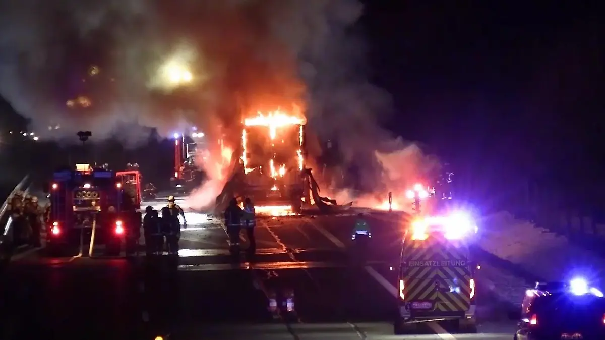 Ein brennender Lkw steht am rechten Rand einer dreispurigen Autobahn und brennt. Vier Feuerwehrfahrzeuge stehen um den brennenden Lkw herum, Feuerwehrkräfte versuchen den Brand zu löschen.