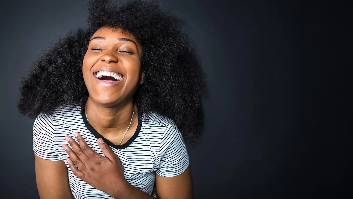 Cheerful young woman with long afro hairstyle against black background. Female is laughing with eyes closed. She is in striped t-shirt.
