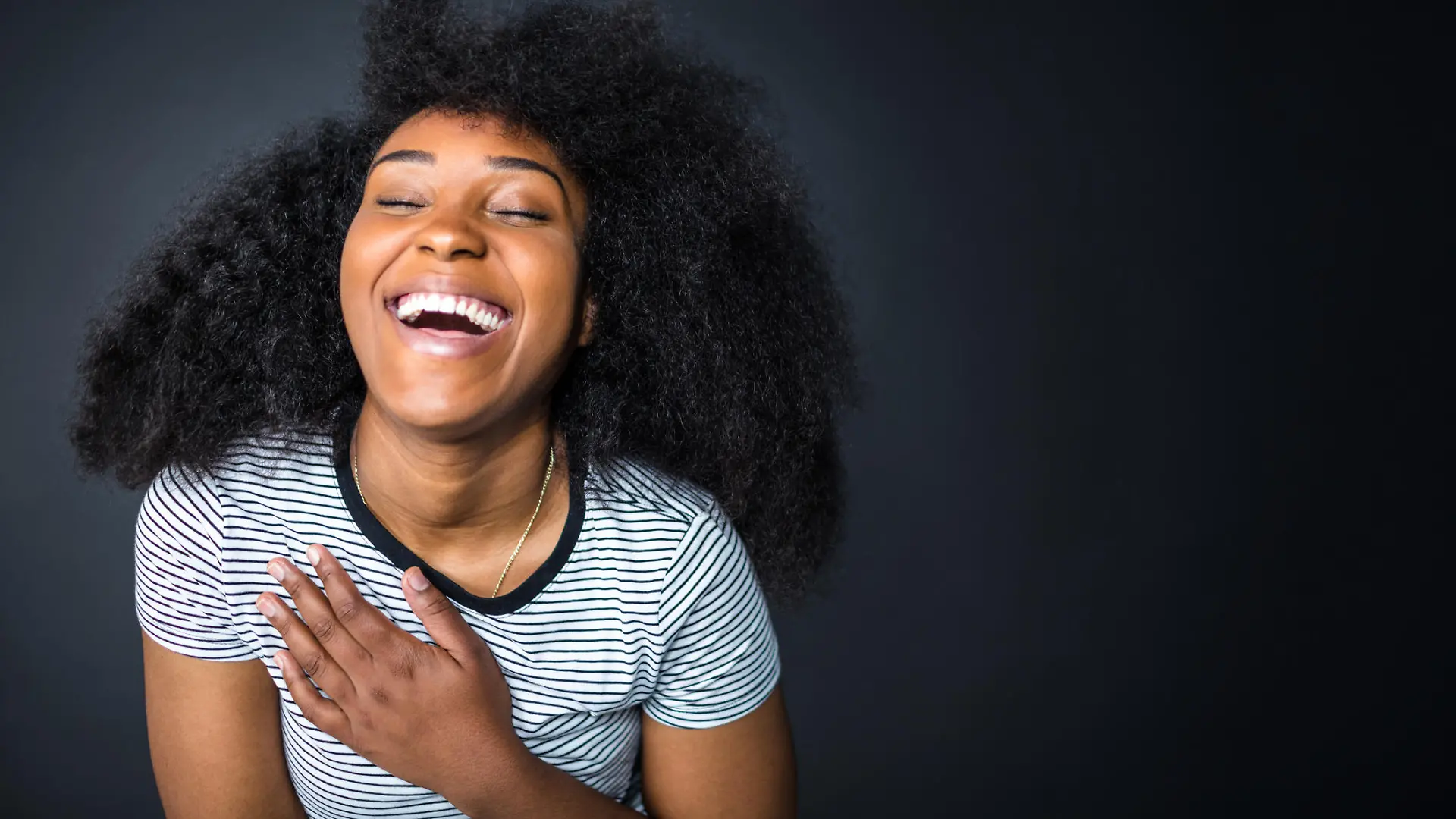 Cheerful young woman with long afro hairstyle against black background. Female is laughing with eyes closed. She is in striped t-shirt.