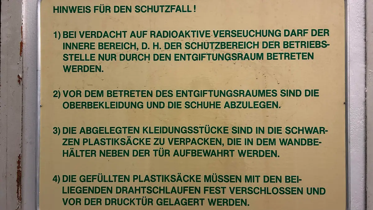 Ein gelbes Schild hängt an einer weißen Wand an der die Farbe absplittert. Auf dem Schild steht in grüner Schrift, wie Mitarbeiter sich im Falle einer radioaktiven Verseuchung verhalten sollten.