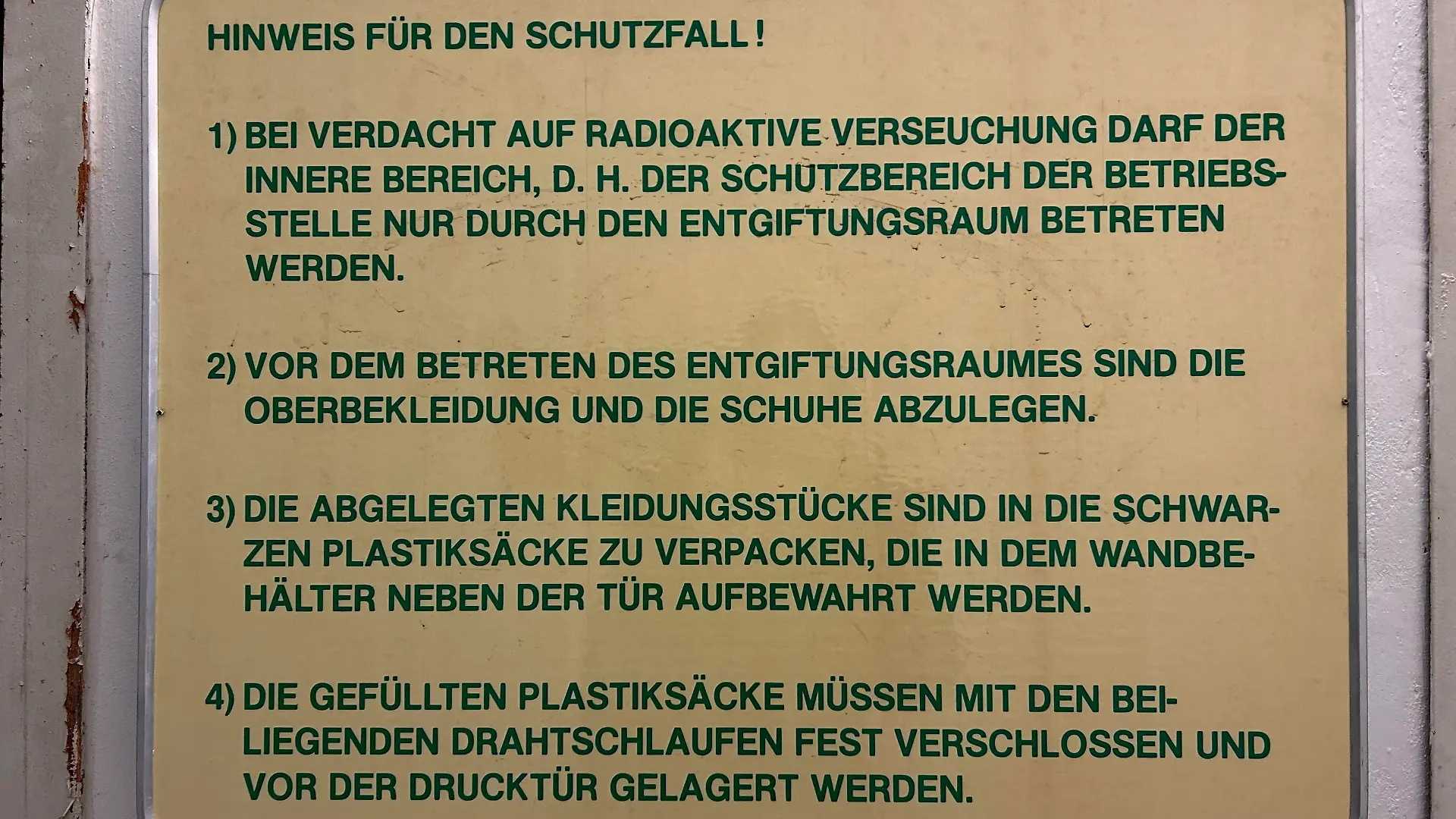 Ein gelbes Schild hängt an einer weißen Wand an der die Farbe absplittert. Auf dem Schild steht in grüner Schrift, wie Mitarbeiter sich im Falle einer radioaktiven Verseuchung verhalten sollten.