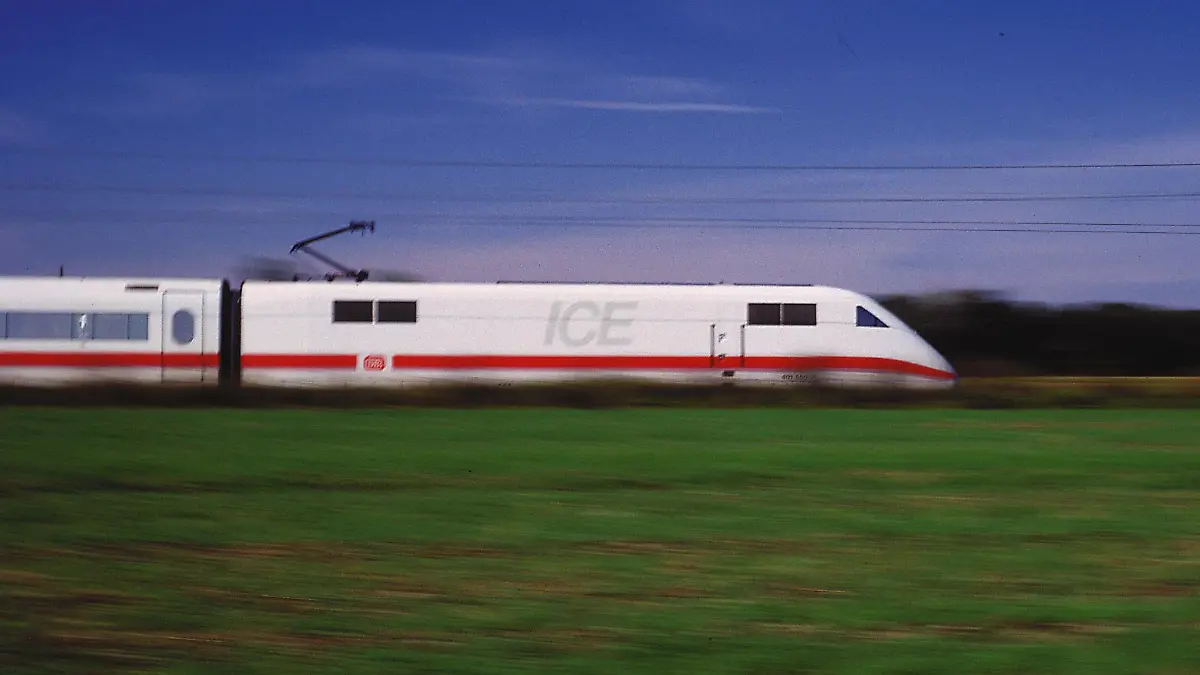 Ein fahrender ICE Zug durch eine Wiese. Im Hintergrund ist Wald und blauer Himmel.