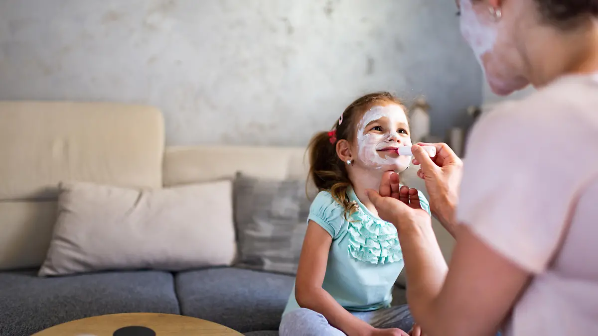 Beautiful woman applying facial mask to daughter’s face. Virtual spa day during COVID-19 isolation.