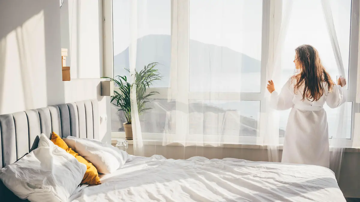 Woman in a bathrobe opening curtains in room at sunrise.