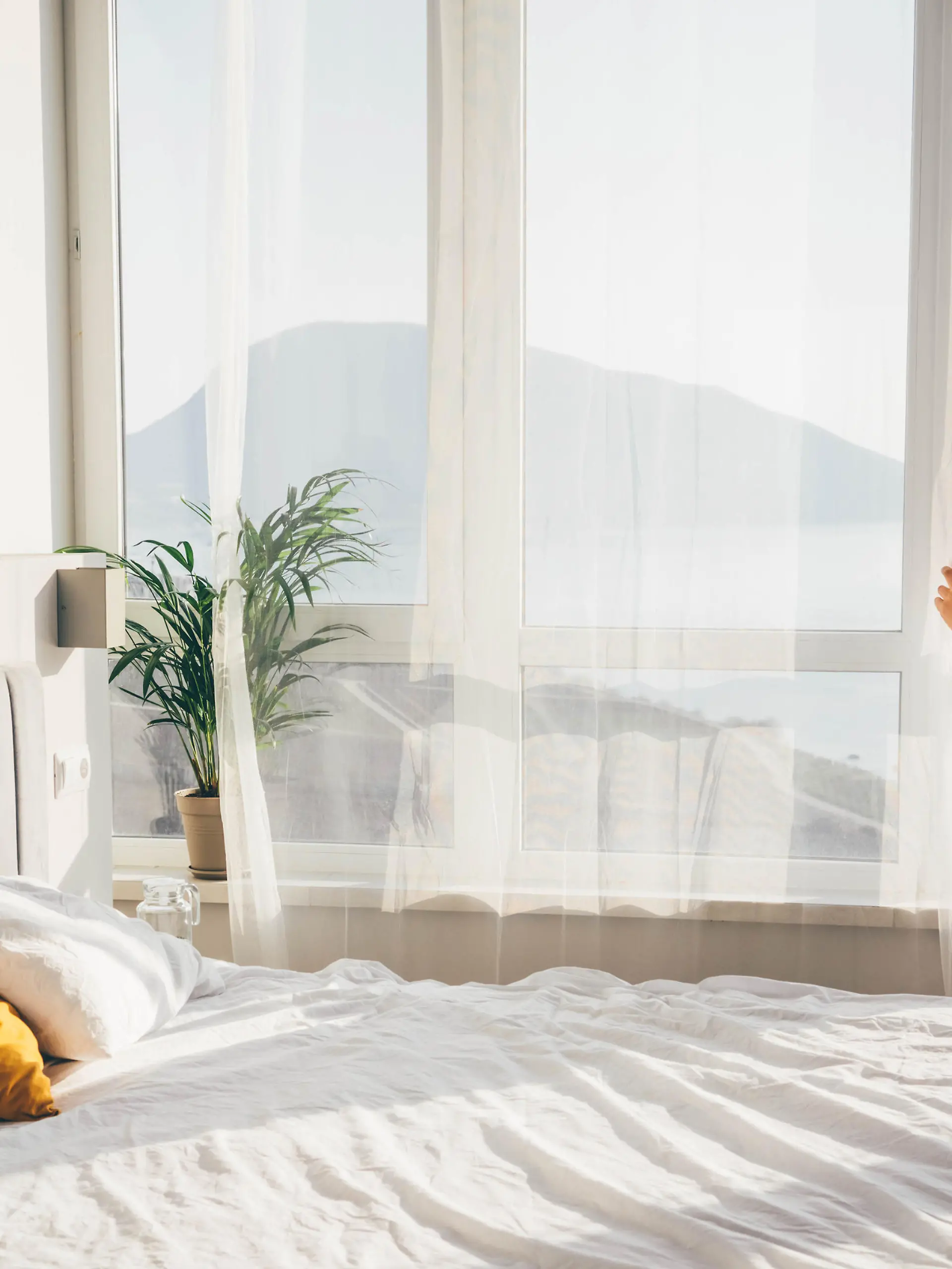 Woman in a bathrobe opening curtains in room at sunrise.
