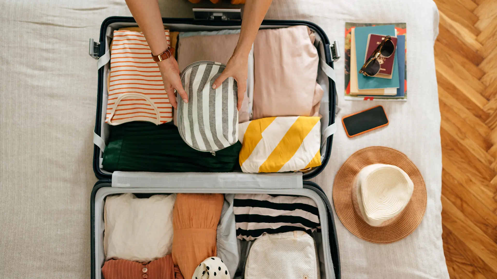 Shot from above of an anonymous woman packing things in her suitcase on the bed. She is holding and putting a grey and white stripped cosmetics bag in. There is her mobile phone, books and sunglasses on the bed.