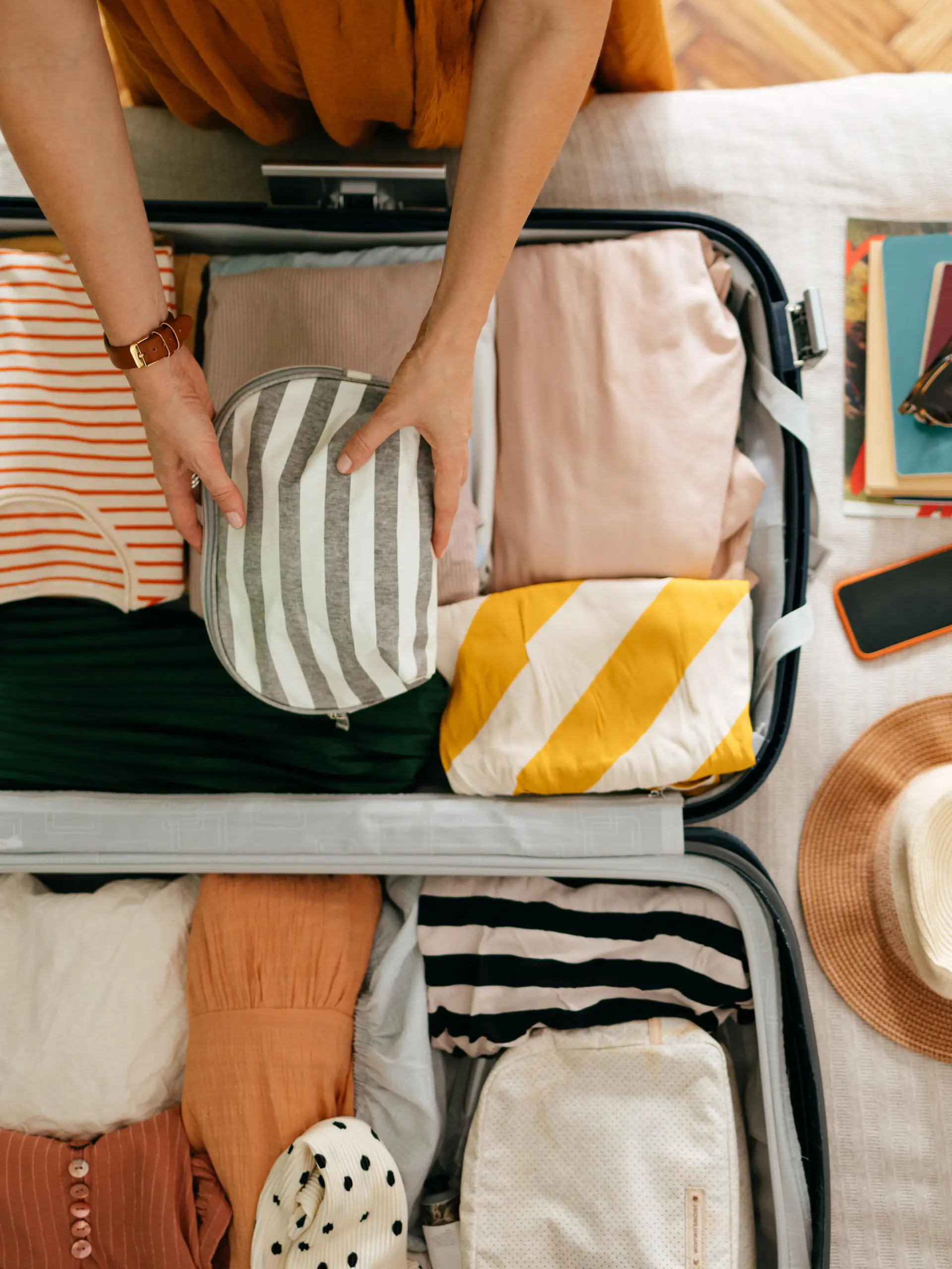 Shot from above of an anonymous woman packing things in her suitcase on the bed. She is holding and putting a grey and white stripped cosmetics bag in. There is her mobile phone, books and sunglasses on the bed.