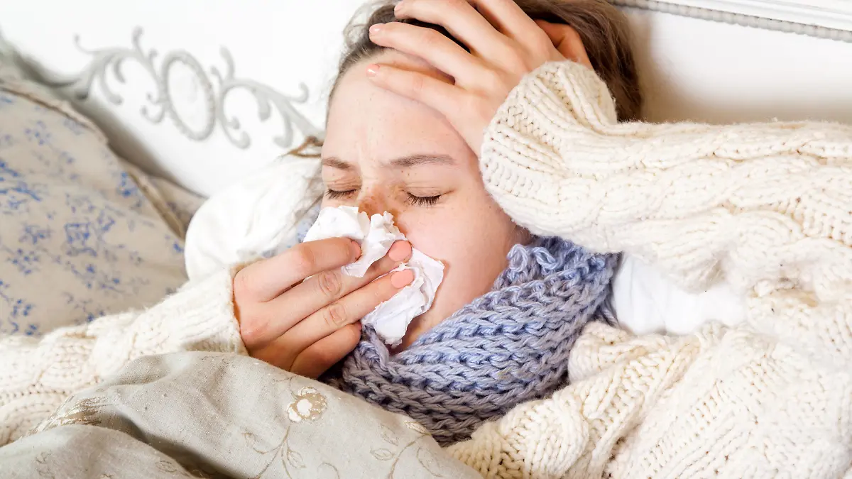 Flu. Closeup image of frustrated sick woman lying in bed in thick blue scarf holding tissue by her nose and touching her head and blowing her nose with closed eyes. || Modellfreigabe vorhanden