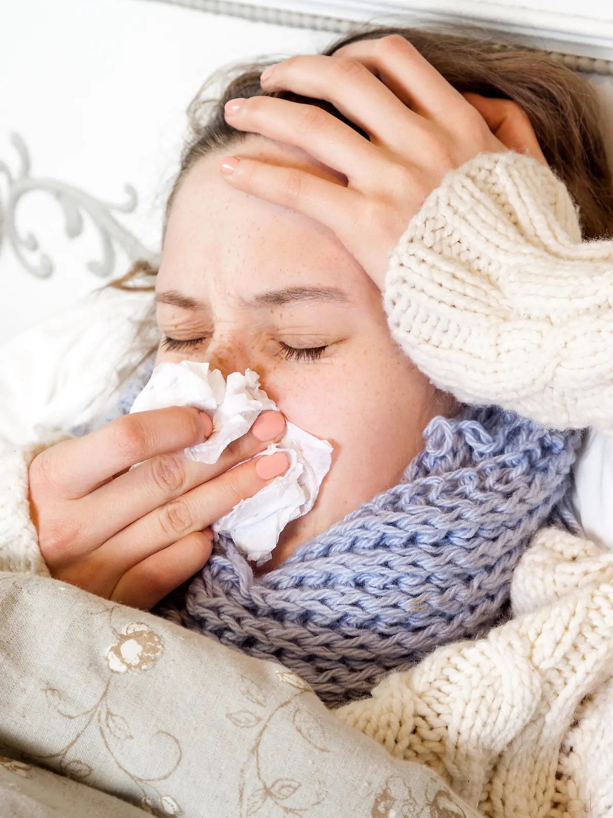 Flu. Closeup image of frustrated sick woman lying in bed in thick blue scarf holding tissue by her nose and touching her head and blowing her nose with closed eyes. || Modellfreigabe vorhanden