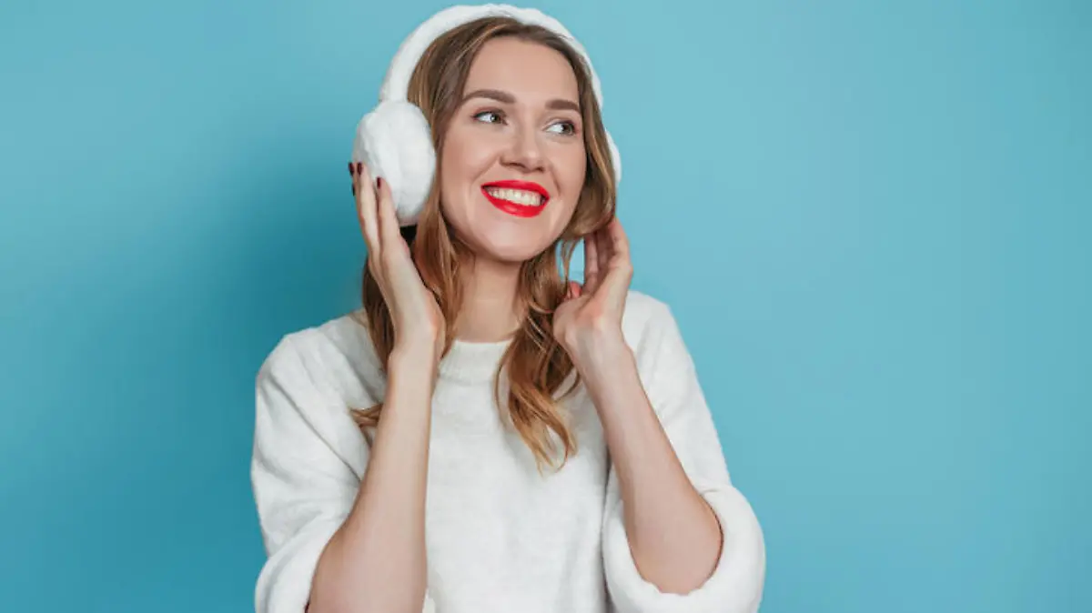 Happy smiling young caucasian woman with fur headphones and white sweater smiling and looking at copy space isolated on blue studio background