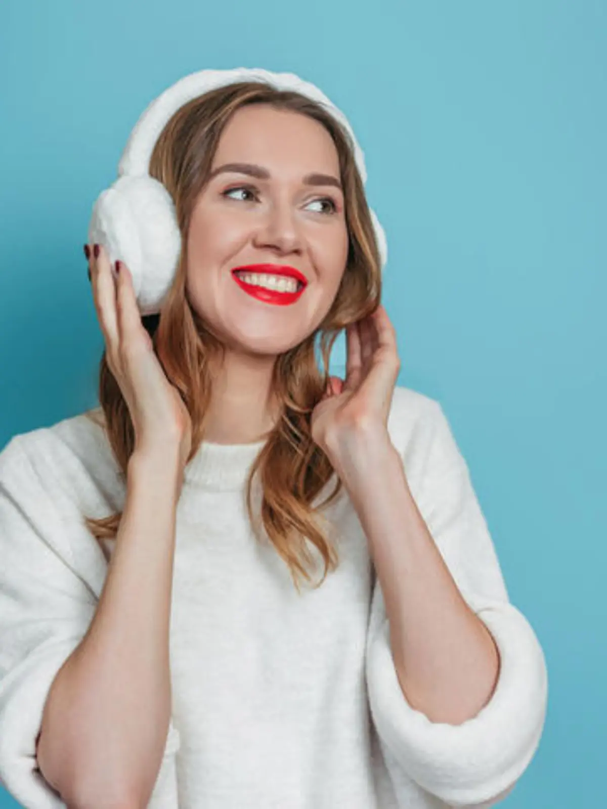 Happy smiling young caucasian woman with fur headphones and white sweater smiling and looking at copy space isolated on blue studio background