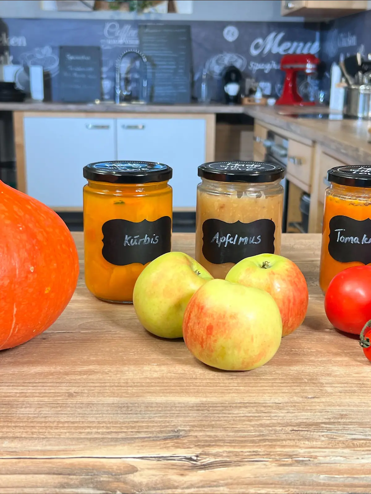 Auf diesem Bild sehen Sie drei fertige Einweckgläser mit der Aufschrift Kürbis, Apfelmus und Tomatensoße. Vor den Gläsern liegen die jeweiligen Gemüse und Obstsorten diese eingekocht wurden. Im Hintergrund sehen Sie eine Küche.