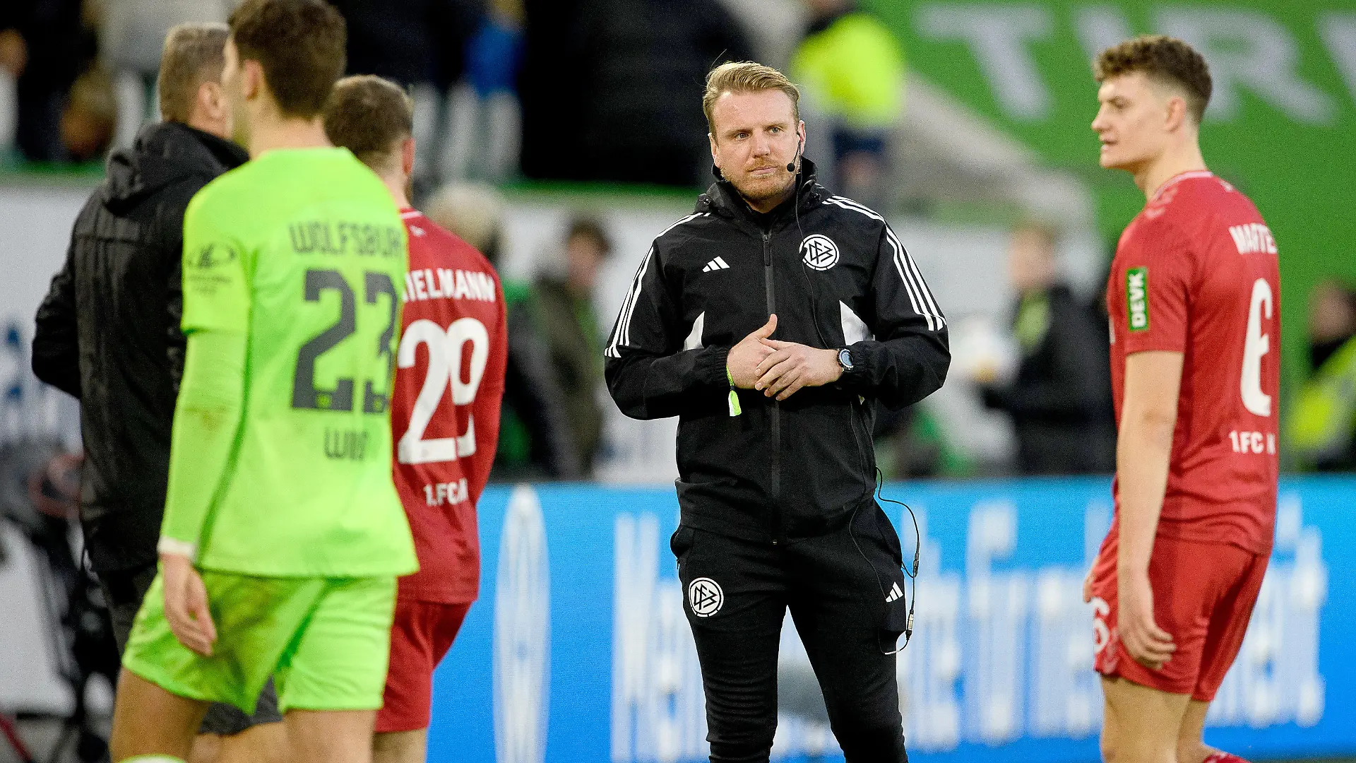 27.01.2024, Niedersachsen, Wolfsburg: Fußball, Bundesliga, VfL Wolfsburg - 1. FC Köln, 19. Spieltag, Volkswagen Arena. Tobias Krull (2.v.r.) übernimmt als ehrenamtlicher Schiedsrichter nach einer Schiedsrichter-Assistenverletzung die Position des vierten Offiziellen. Der frühere Wolfsburger Nachwuchsspieler Tobias Krull ist Torwart und Sportlicher Leiter beim MTV Gifhorn in der Fußball-Landesliga. Er meldete sich während der ersten Halbzeit, als der Stadionsprecher per Lautsprecher-Durchsage jemanden mit Schiedsrichter-Lizenz im Publikum suchte. Es wurde ein Ersatz für das vierte Mitglied des Schiedsrichter-Teams gesucht, das an der Seitenlinie zwischen den beiden Trainern steht. Foto: Swen Pförtner/dpa - WICHTIGER HINWEIS: Gemäß den Vorgaben der DFL Deutsche Fußball Liga bzw. des DFB Deutscher Fußball-Bund ist es untersagt, in dem Stadion und/oder vom Spiel angefertigte Fotoaufnahmen in Form von Sequenzbildern und/oder videoähnlichen Fotostrecken zu verwerten bzw. verwerten zu lassen. +++ dpa-Bildfunk +++