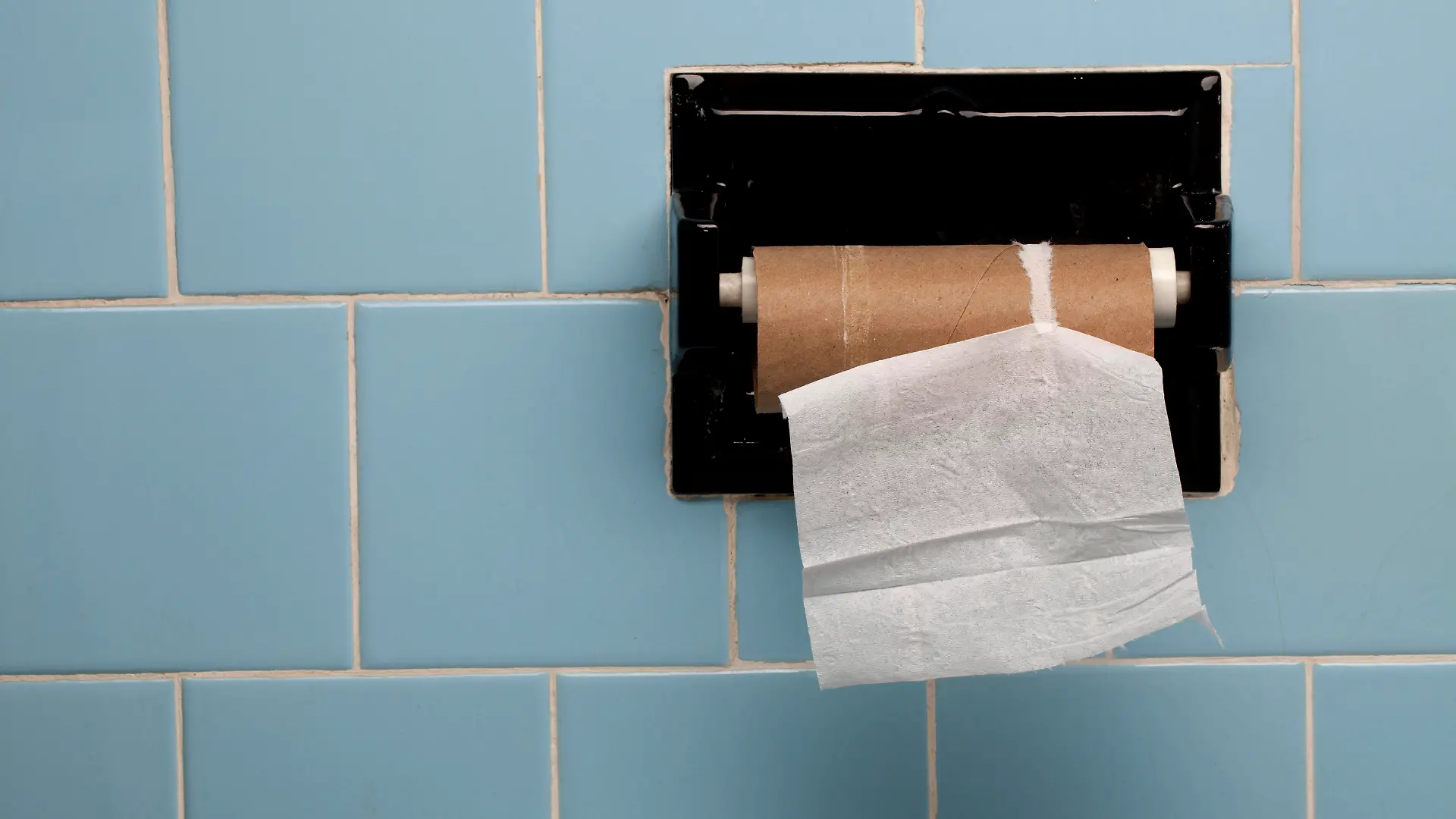 Vintage blue tile on a bathroom wall with only one piece of toilet paper left on the roll.