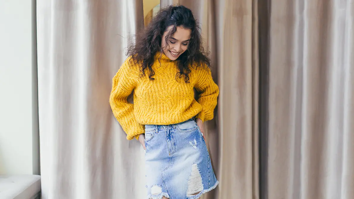 Portrait of a young beautiful woman girl dressed and posing in new clothes in a yellow warm sweater and denim skirt in a clothing store on a background of brown curtains near the fitting room, posing, showing, bragging, looking at the camera, smiling